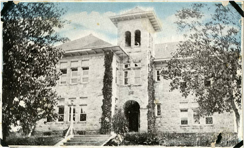 A postcard of the Cantril Schools east facade, located at 312 Cantril Street., Castle Rocks Cantril. The two-story rhyolite structure has a bell tower and is made of this material. Double-hung windows with transom lights are present throughout the facility. The entrance is through the arched entry under the bell tower and is accessed via steps. On either side of the building are trees. Its colored up in the sky 1910-1930.