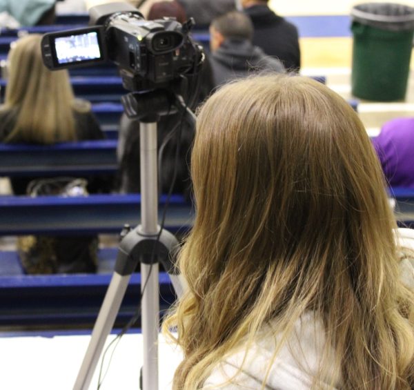 A student sits in the stands at a high school basketball game with a video camera.