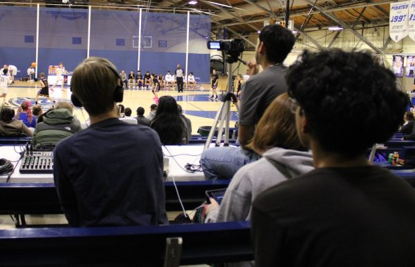 Students sit in the stands at a high school basketball game with a video camera and audio equipment.
